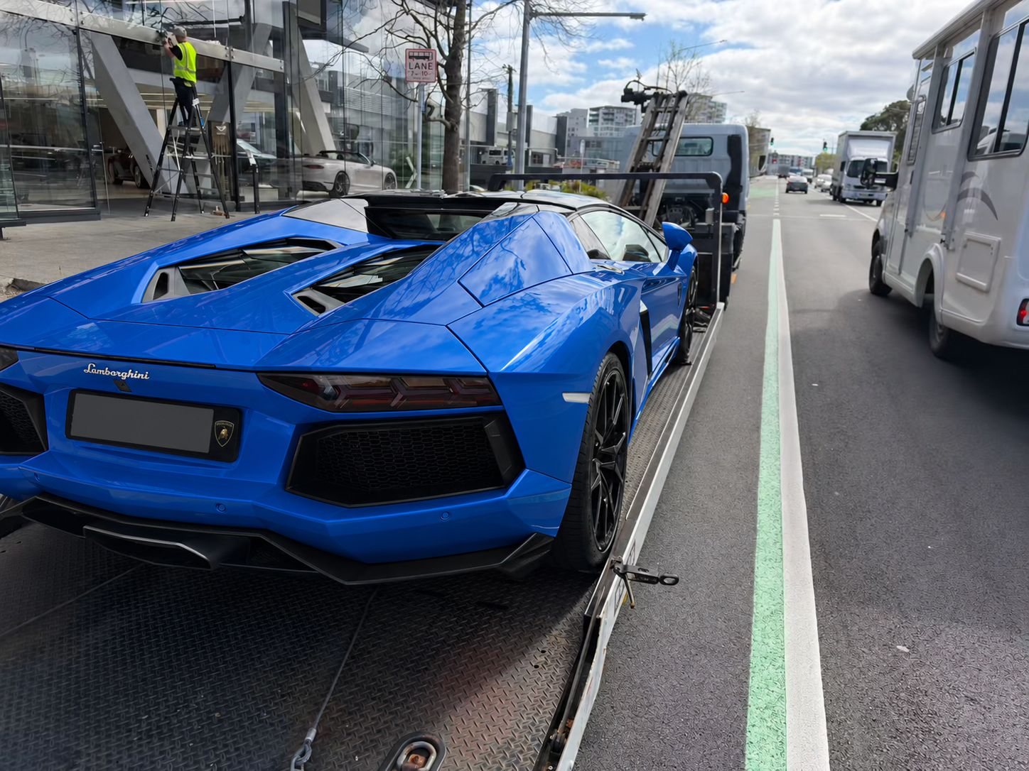 Rear of the Lamborghini on a flatbed, showing the diffuser and a fully covered registration plate; body colour edited to blue.